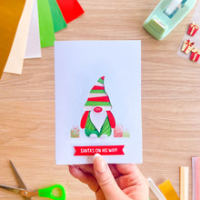 Load image into Gallery viewer, A hand holds a square greeting card featuring a Christmas gonk with the text 'SANTA'S ON HIS WAY!' on a light wood desk, surrounded by craft supplies.