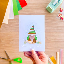 Load image into Gallery viewer, A hand holds a square handmade greeting card featuring an iris fold gonk with a green and gold striped hat, surrounded by craft supplies on a wooden desk.