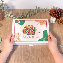 Load image into Gallery viewer, Person holding a gift box with a colourful hedgehog design and 'Enjoy the Moment' text on a wooden surface with decorative elements.