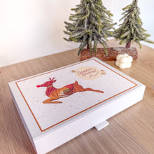 Load image into Gallery viewer, White box with geometric reindeer design on a wooden surface with Christmas trees in the background
