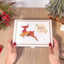 Load image into Gallery viewer, Person holding a Christmas gift box with a reindeer design on a wooden surface with festive decorations.