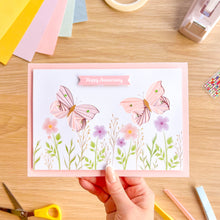 Load image into Gallery viewer, Handmade card with pink iris folding butterflies and lilac flowers on a wooden table with crafting supplies.