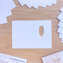 Load image into Gallery viewer, Hand holding a scored card blank with a champagne glass cut-out with printed gold stem on a wooden surface