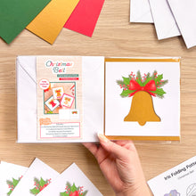 Load image into Gallery viewer, A hand holds a Christmas Bell card aperture pack on a light wood tabletop. The package shows a card design with a gold bell, green holly leaves, red berries, and a red bow.