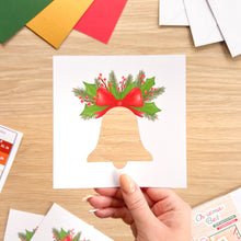 Load image into Gallery viewer, A hand holds a white square card cut-out featuring a Christmas bell design with green holly leaves, red berries, and a red bow. The card is photographed on a light wood tabletop with other craft or stationery items partially visible around it.