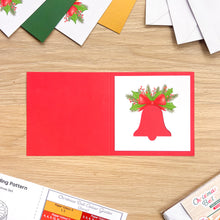 Load image into Gallery viewer, A red greeting card laid flat on a light wood surface, shows a white cut-out of a Christmas bell with green holly and red bow illustration. Other cards and envelopes are partially visible around the edges.