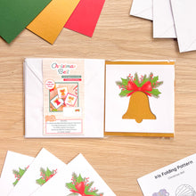 Load image into Gallery viewer, A Christmas bell card aperture pack displayed on a light wood tabletop, showing the printed package label, the card cut-out on a gold blank card with a red bow, and green holly, plus several folded cards and envelopes around it.