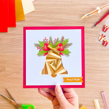 Load image into Gallery viewer, A hand holds a square Christmas greeting card with a gold geometric bell, green holly leaves, red berries, and a red border on a light wood desk with craft supplies scattered around.
