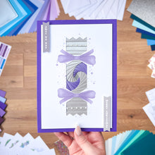 Load image into Gallery viewer, Hand holding a card with purple and silver Cracker design on a wooden surface with craft materials.