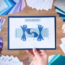 Load image into Gallery viewer, Hand holding a Cracker card with blue ribbons and text 'All is Calm, All is Bright' against a colorful background of craft materials.