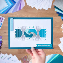 Load image into Gallery viewer, Hand holding a decorative Cracker card with blue and white design on a wooden surface with craft materials.