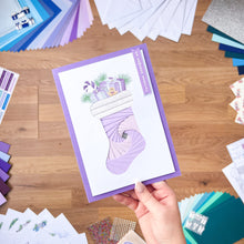 Load image into Gallery viewer, Hand holding a Stocking greeting card with a purple border on a wooden surface with various craft materials.