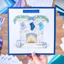 Load image into Gallery viewer, Christmas card with stockings by a fireplace held in front of craft materials on a wooden surface.