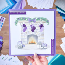 Load image into Gallery viewer, Hand holding a Christmas card with stockings by a fireplace on a wooden surface.