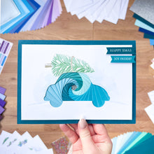 Load image into Gallery viewer, Hand holding a Christmas card with a car in a blue iris fold spiral design and festive text on a wooden surface.