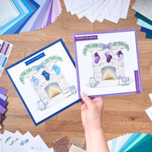 Load image into Gallery viewer, Hand holding a card with a festive design of stockings and a fireplace, surrounded by craft materials on a wooden surface.