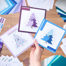 Load image into Gallery viewer, Hand holding three Christmas cards with geometric tree design, surrounded by craft materials on a wooden surface.