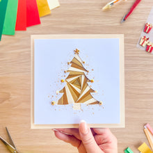 Load image into Gallery viewer, A hand holds a square greeting card featuring an iris fold Christmas tree design on a speckled background. The card is photographed from above on a light wood desk surrounded by craft supplies, including colored paper sheets, scissors, and small gift boxes.