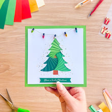 Load image into Gallery viewer, A hand holds a square handmade Christmas card featuring a green layered tree design with colorful string lights and the text 'Have a lovely Christmas' on a light wood background with craft supplies.