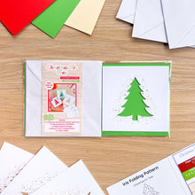 Load image into Gallery viewer, An iris folding card aperture pack on a light wood surface, showing a green Christmas tree design on the right panel and the pack label on the left. White envelopes and colored paper sheets are arranged around it on a wooden tabletop.