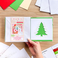 Load image into Gallery viewer, A hand holds a Christmas Fir Tree card aperture pack on a wooden table, showing a green tree cut-out with gold green glitter accents. Pack contents are scattered around the table.