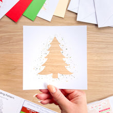 Load image into Gallery viewer, A hand holds a square white card featuring a gold glitter Christmas tree design on a light wood tabletop. Colorful paper sheets and envelopes are scattered in the background.