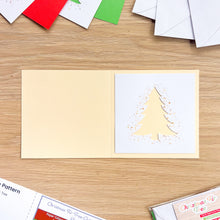 Load image into Gallery viewer, An open handmade greeting card on a light wood tabletop. The left panel is a warm cream color, and the right panel features a layered cream Christmas tree silhouette with gold glitter speckles on a white backing.