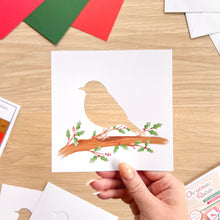 Load image into Gallery viewer, A hand holds a square white greeting card cut-out featuring a bird silhouette perched on a decorated branch with green holly leaves and red berries, set on a light wood tabletop with other cards and paper pieces around it.
