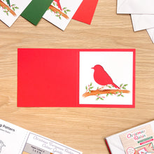 Load image into Gallery viewer, A red greeting card blank with a white embellished bird cut-out featuring a bird perched on a branch, displayed on a light wood surface with craft pattern sheets and envelopes around it.