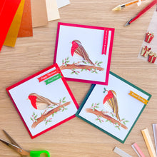 Load image into Gallery viewer, Three handmade greeting cards with bird illustrations are arranged on a wooden craft table alongside scissors, colored paper, and small decorative items. The cards feature red, white, and green borders with a bird design and holiday text.