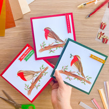 Load image into Gallery viewer, Three handmade greeting cards featuring a red-breasted bird on a branch with holly leaves, arranged on a wooden craft table. A hand is holding one green-bordered card, while two more cards with red and dark red borders are placed above it.