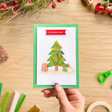 Load image into Gallery viewer, A hand holds a handmade Christmas card featuring a colourful iris fold Christmas tree and gift boxes, set against a wooden desk with holiday decorations.