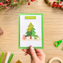 Load image into Gallery viewer, A hand holds a handmade Christmas card featuring a green layered paper Christmas tree and presents under a 'Merry Christmas' banner, set against a wooden craft table with scissors and ribbon.