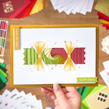 Load image into Gallery viewer, Hand holding a Christmas Cracker card with colorful ribbons and 'Happy Xmas' text on a wooden surface.