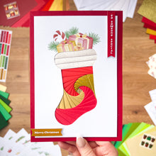 Load image into Gallery viewer, Christmas card with a colorful stocking design held by a hand on a wooden surface.