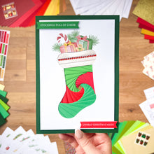 Load image into Gallery viewer, Hand holding a Christmas card with a colorful stocking design on a wooden surface.