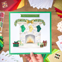Load image into Gallery viewer, Hand holding a Christmas card with stockings and fireplace design on a wooden surface.