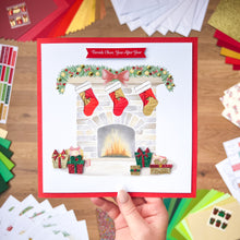 Load image into Gallery viewer, Hand holding a Christmas card with stockings and a fireplace design, surrounded by craft supplies.