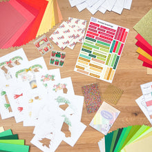 Load image into Gallery viewer, Assorted craft materials from the Christmas at home kit including paper, stickers, and cards on a wooden surface.