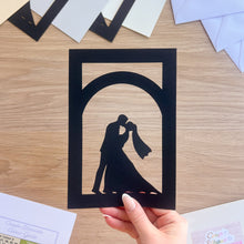 Load image into Gallery viewer, Hand holding a black paper cutout of a couple kissing against a wooden background