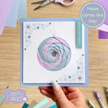 Load image into Gallery viewer, Handmade card with decorative design held by a hand, surrounded by crafting materials on a wooden surface.