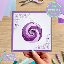 Load image into Gallery viewer, Hand holding a card with a purple bauble design on a wooden table with crafting supplies.