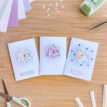 Load image into Gallery viewer, Three handmade greeting cards with geometric layered paper cutouts are arranged on a wooden desk alongside crafting tools like scissors, tweezers, and paper scraps.