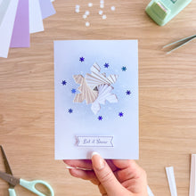 Load image into Gallery viewer, A handmade greeting card held by a hand, featuring a layered paper snowflake design with blue and beige tones on a light blue background, with the text 'Let it Snow' at the bottom. The card is photographed on a wooden desk with crafting tools like scissors, paper, and a tape dispenser visible around it.