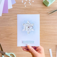 Load image into Gallery viewer, A hand holds a light blue greeting card featuring an iris fold snowflake design in the center and a small blue banner reading 'Season's Greetings' at the bottom, set against a wooden desk with crafting supplies.