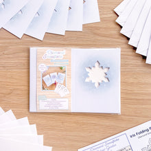 Load image into Gallery viewer, A packaged diamond snowflake card aperture pack lies on a light wood tabletop, surrounded by scattered white paper envelopes, card cut-outs, paper sheets and iris folding pattern. The package front shows a snowflake cutout window and card examples.
