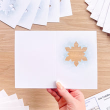 Load image into Gallery viewer, A hand holds an open white folding card on a light wood table. The right panel shows a printed snowflake design with a soft blue watercolor wash.