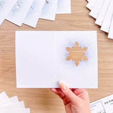 Load image into Gallery viewer, A hand holds an open white greeting card with a cutout snowflake design on the right side, set on a light wood tabletop with other cards scattered around.