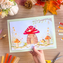 Load image into Gallery viewer, A hand holds a colorful printed card featuring a whimsical iris fold toadstool house, set against a wooden tabletop with craft supplies and autumn leaves around it.