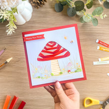 Load image into Gallery viewer, A handmade square greeting card with a red border and a red-and-white iris fold mushroom design, held by a hand over a wooden table with craft supplies and flowers.